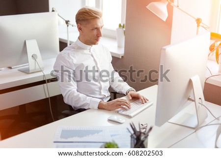 Young blond man sitting at the computer and typing in the office. Horizontal indoors shot.