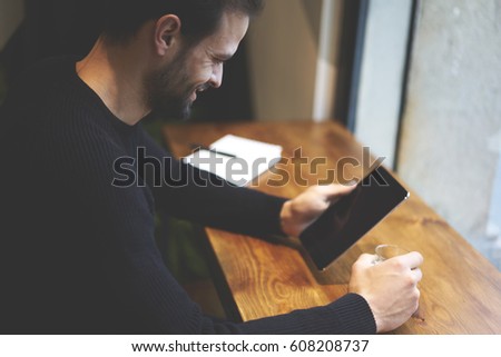 Cropped image positive handsome freelancer dressed in style black sweater watching funny movie on digital tablet using free internet connection and enjoying morning coffee sitting in cozy cafeteria