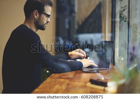 Handsome caucasian student writing coursework on modern laptop using 3G internet connection while sitting in cafe. Hipster guy working distantly via computer with actualised software for modern device