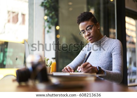 Concentrated afro american experienced woman journalist in glasses creating article working in coffee shop, skilled young female graphic designer making sketch in notebook for new startup project  Royalty-Free Stock Photo #608213636