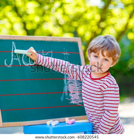 Adorable funny little kid boy at blackboard practicing writing letters, outdoor school or nursery. Child having fun with learning. Back to school concept.