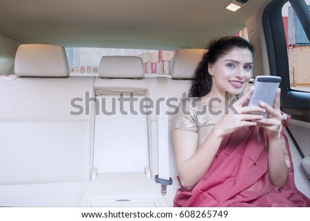 Picture of a successful Indian female importer sitting in the car while using mobile phone and wearing saree clothes with container on the background