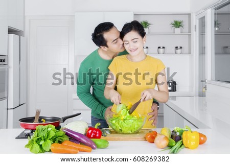 Picture of a young man kissing his wife while making vegetables salad in the kitchen at home