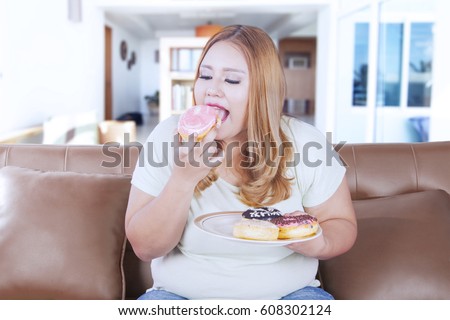 Picture of a blonde obesity woman sitting on the sofa while eating donuts at home