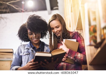 Two multiethnic young female friends enjoying study together in a cafe with library books, laughing and talking. African and caucasian hipster student girls brainstorming together