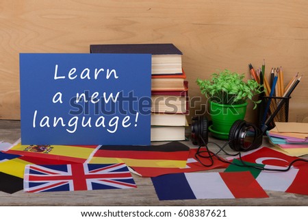 Learning languages concept - paper with text "Learn a new language!", flags, books, headphones, pencils on wooden background Royalty-Free Stock Photo #608387621