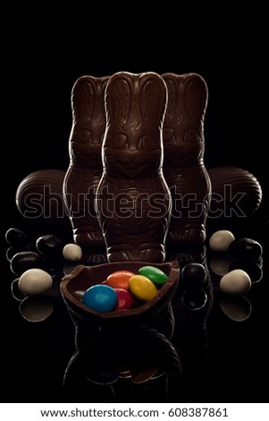 Three Chocolate easter bunny and eggs with black and white and colored sweets close-up macro in dark, on black background, with beautiful white outline. concept of holiday and happy Easter.