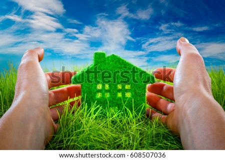 Eco House in green grass protected by the human hands on blue sky background.