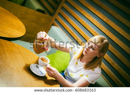 Young blonde woman wearing white T-shirt with print, girl pours a fruit drink from a glass jug, into a white cup, sitting at a table in a cafe, background stylized Wood texture lit up with warm light