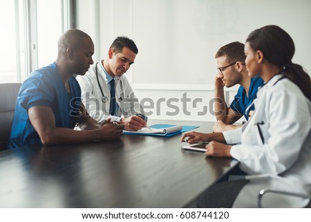 Multiracial medical team having a meeting with doctors in white lab coats and surgical scrubs seated at a table discussing a patients records Royalty-Free Stock Photo #608744120