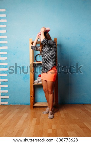 Portrait of a young attractive woman in blue room on wooden cupboard background posing. hipster woman holding book on head. balancing. Education concept photo, lifestyle. training the posture