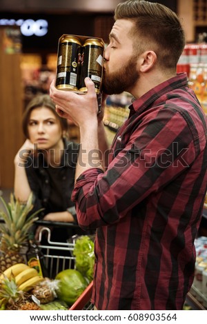 Picture of young loving couple in supermarket with shopping trolley choosing products. Dissatisfied woman looking at man holding beer.
