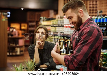 Picture of young loving couple in supermarket with shopping trolley choosing products. Dissatisfied woman looking at man holding beer.