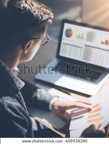 Young finance market analyst in eyeglasses working at sunny office on laptop while sitting at wooden table.Businessman analyze document in his hands.Graphs and diagramm on notebook screen.Vertical
