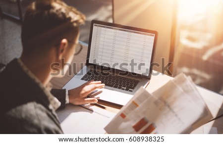 Young banking finance analyst in eyeglasses working at sunny office on laptop while sitting at wooden table.Businessman analyze stock reports on notebook screen.Blurred background,horizontal Royalty-Free Stock Photo #608938325