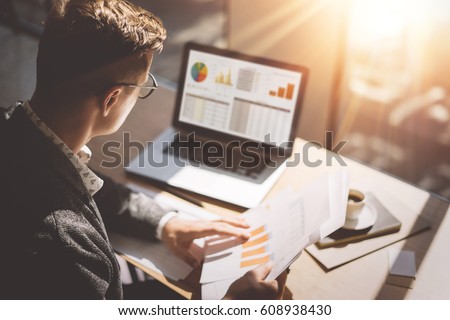Young finance market analyst in eyeglasses working at sunny office on laptop while sitting at wooden table.Businessman analyze document in his hands.Graphs and diagramm on notebook screen.Blurred Royalty-Free Stock Photo #608938430