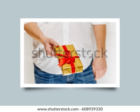 Male hand holding a gift box. Present wrapped with ribbon and bow. Christmas or birthday golden package. Man in white shirt. Photo frame design with shadow.