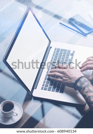 Adult tattooed coworker working on laptop with white blank computer screen at sunny office.Businessman typing on notebook keyboard.Vertical mockup,blurred background