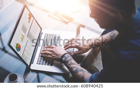 Adult tattooed coworker using laptop while working at sunny office.Graphs and diagramm on notebook screen.Blurred background.Horizontal. Visual effects, flares