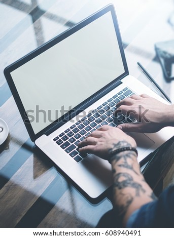 Young tattooed coworker working on laptop with white empty computer screen at sunny office.Businessman typing on notebook keyboard.Vertical mockup,blurred background