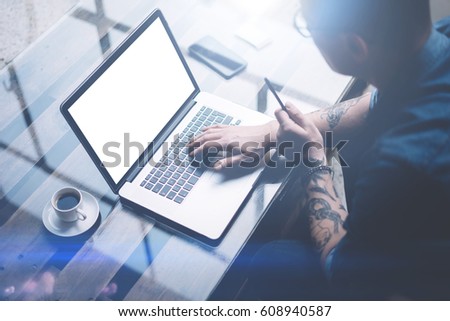 Adult tattooed businessman working on mobile computer at sunny office.Businessman typing on notebook keyboard.White blank laptop screen.Horizontal mockup,blurred background