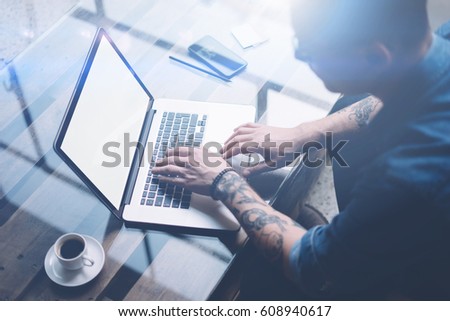 Adult tattooed coworker working on laptop with white blank computer screen at sunny office.Businessman typing on notebook keyboard.Horizontal mockup,blurred background