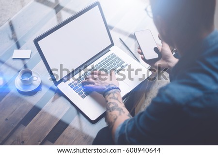 Adult tattooed coworker holding smartphone on hand and using laptop while working at sunny office.White blank notebook screen.Blurred background.Horizontal mockup