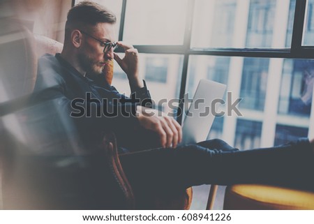 Bearded elegant businessman working at the modern loft office.Man sitting in vintage chair,holding in hand glass of water,using contemporary notebook.Panoramic window on blurred background.Horizontal.
