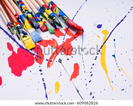 Close-up shot of vivid splashes of paint on white canvas and colorful painting brushes near. 