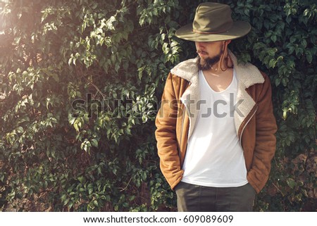 Photo of a young bearded cowboy. Man in a white t-shirt, hat and jacket. Plants background. Horizontal mock up.