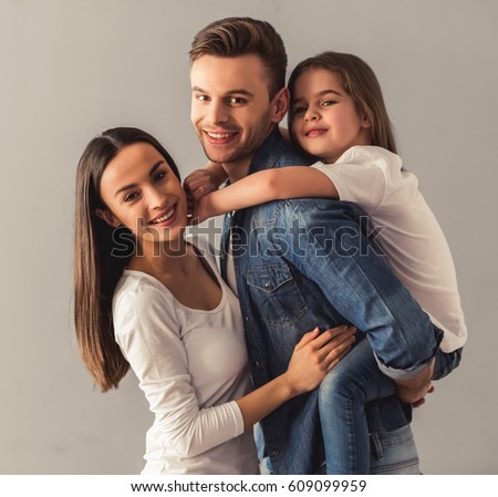 Charming little girl and her beautiful young parents are looking at camera and smiling, on gray background