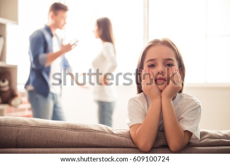 Sad little girl is looking at camera while her parents are arguing in the background Royalty-Free Stock Photo #609100724