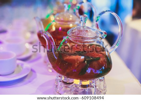 Beautiful warm picture of transparent teapot kettle with tasty green black tea with apple, lemon and ginger on a table with candles and with dessert in the background

