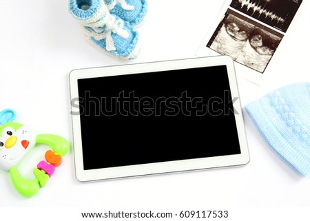 concept of extending family and expecting for baby: blank screen tablet pc, first picture ultrasound scan of baby, clothing for newborn - hat and booties, colorful rattle on white background