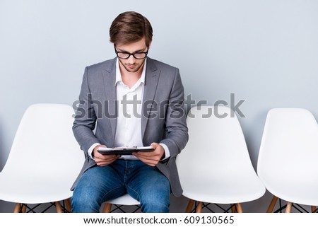 Young man is holding documents while sitting on white chair. He is the last in line for an interview for a new job.