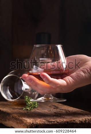 Glass of brandy or cognac in a hand on old oak wooden table. Dark photo.