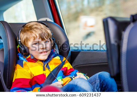 Little blond kid boy watching tv or dvd with headphones during long car driving on family vacations. Leisure for children for long drive. Preschool child sitting in safe car seat.