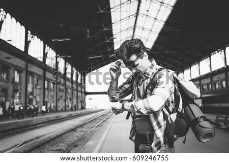 A young man traveling with backpack, waiting for the train, is checking the time on his clock in black and white