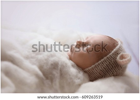 A small baby is dressed in white and covered with a blanket and he is peacefully sleeping during his first photo-shoot.