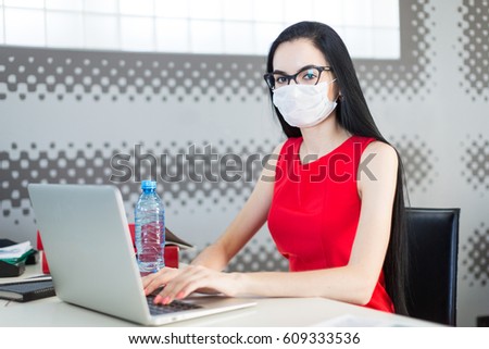 Office on background, pretty, young brunette caucasian business lady in red dress, respirator and glasses sit at the table and work with laptop, water bottle on the table, look at camera