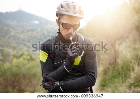 Sports, extreme and active lifestyle. Attractive young European cyclist wearing protective gear holding tube and drinking water out of bladder while resting during intensive workout in mountains