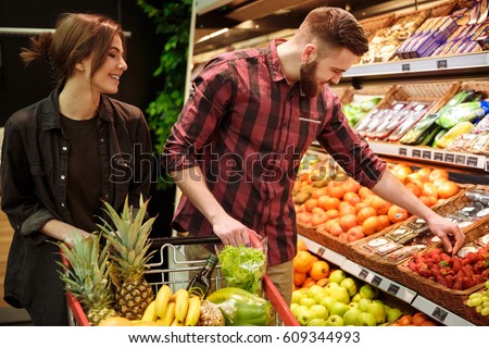 Image of young happy loving couple in supermarket with shopping trolley choosing fruits. Looking aside.