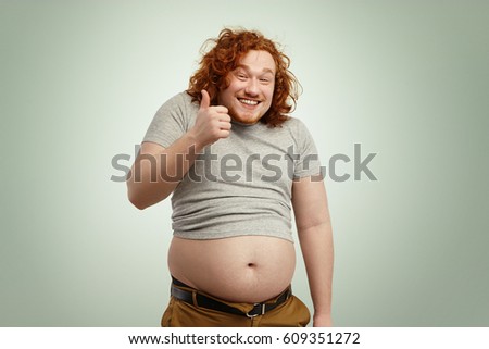 People, unhealthy lifestyle, diet and obesity concept. Fat overweight redhead Caucasian man in undersized t-shirt showing thumps up and smiling happily while his big stomach hanging out of pants