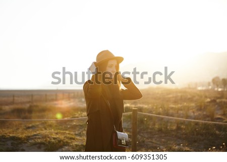 Beautiful picture of young European female dressed in overclothes adjusting brims of her stylish hat while having nice walk alone outdoors at seaside, warm evening sun shining brightly in background