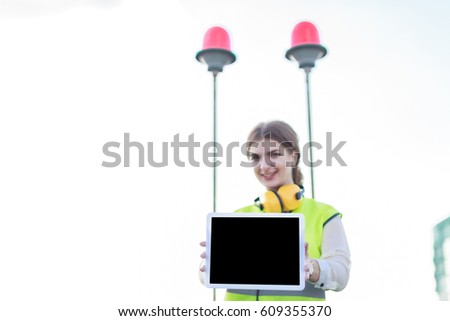 Blurred sky and red signal lights on background, young pretty brunette caucasian woman in green west and yellow earmuffs stand on roof and show empty tablet, look at camera