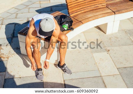 Photo from above of an athletic man taking a break from workout while tying his shoes and sitting on a bench.