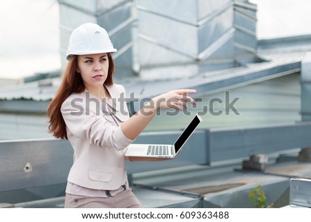 Blurred roof on background, attractive redhead caucasian business lady in beige suit, brown trousers and white helmet sit on the roof and hold  laptop, point at right