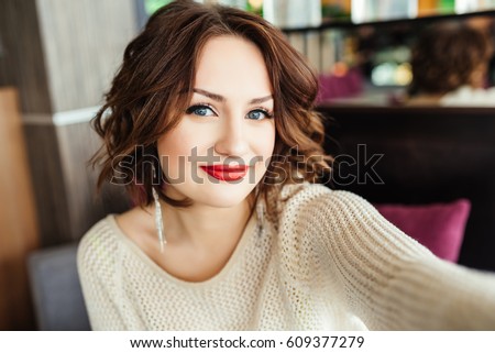 Selfie-portrait of cute brunette girl with short hair sitting at table on terrace in restaurant. She wears yellow sweater and looks happy