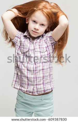 Vertical picture, isolated on grey, adorable, cute little caucasian redhead girl in plaid shirt, bright blue trousers and white boots, look at camera, turned, hands in hair