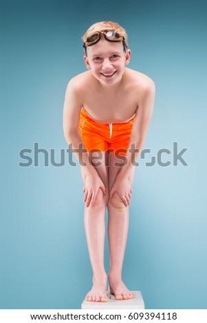 Isolated on grey, vertical picture, teenage blond caucasian boy in orange shorts and swimming glasses, bend to camera